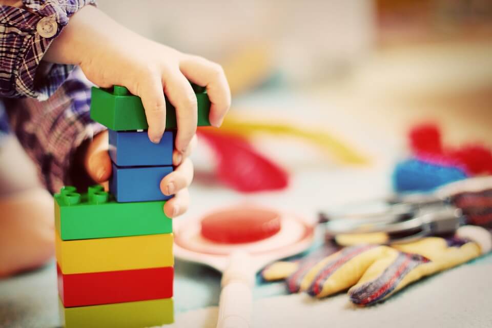 Photograph of young child's hands stacking blue, green, yellow, and red Legos with more toys in the background.