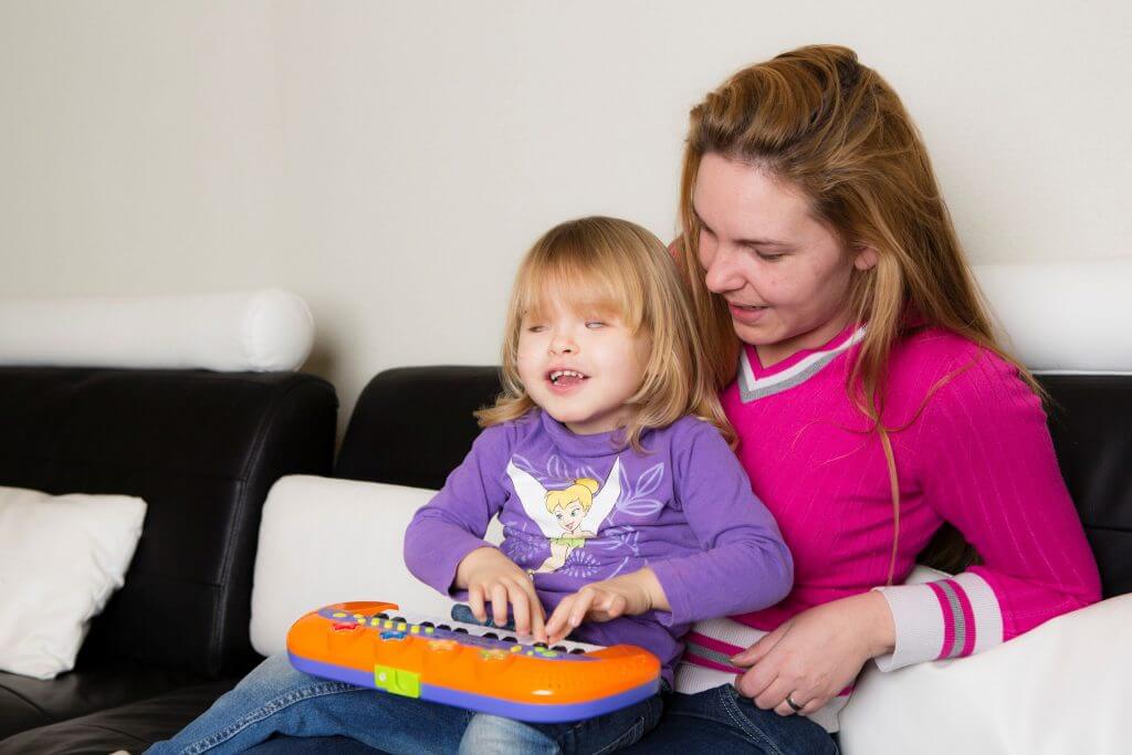 Child with a visual impairment sits on caretaker's lap while playing with a toy piano and smiling.