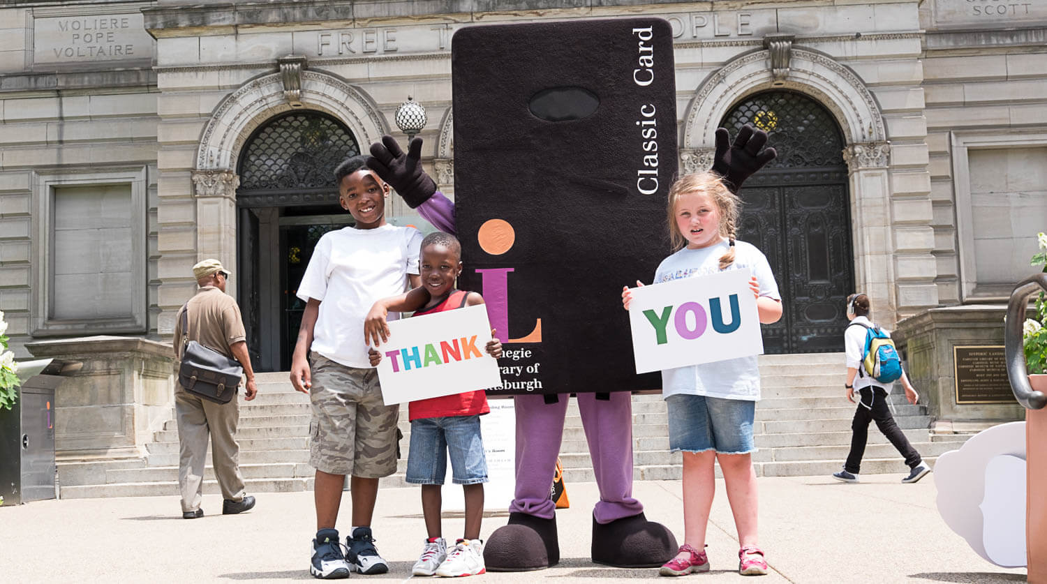 Three children stand in front of the library with Library mascot Andrew Cardnegie while holding thank you signs