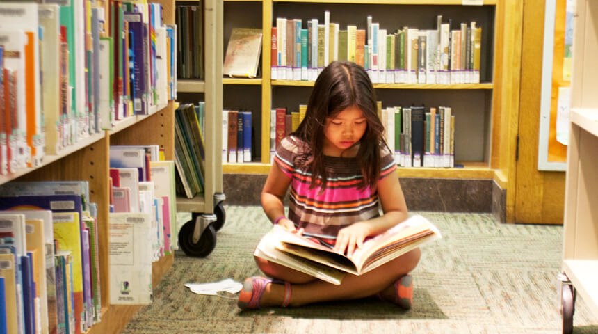 Child sitting cross legged on the floor in the stacks at the library with a book on their lap