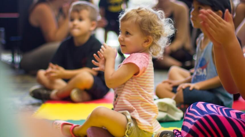 Several children sit on the floor and clap while listening and watching something