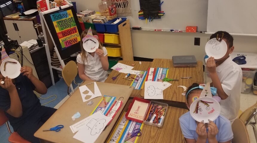 Four third graders hold a paper plate craft up to their faces in a classroom