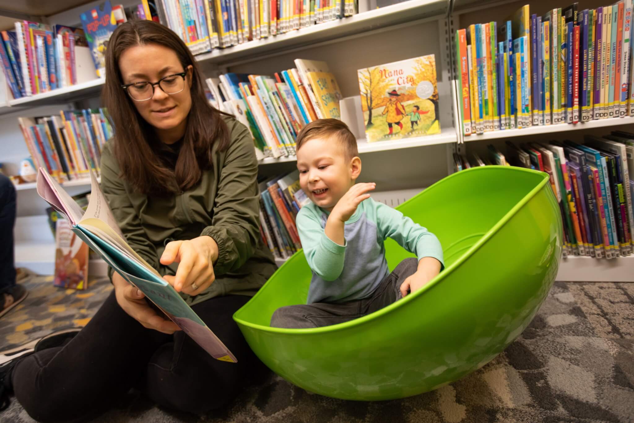 A toddler in a rocking bowl chair is being read to by a caregiver.