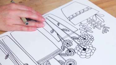A hand holding a pen and resting on a sheet of white paper with a black line drawing of houseplants and potted flowers