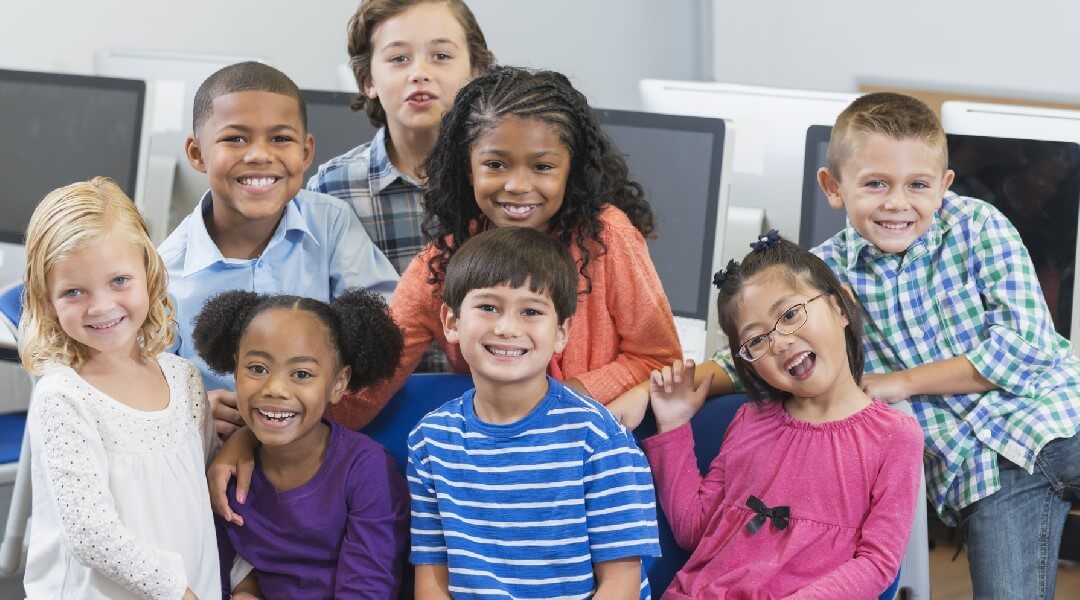 Group of smiling children posing in front of a row of desktop computers.