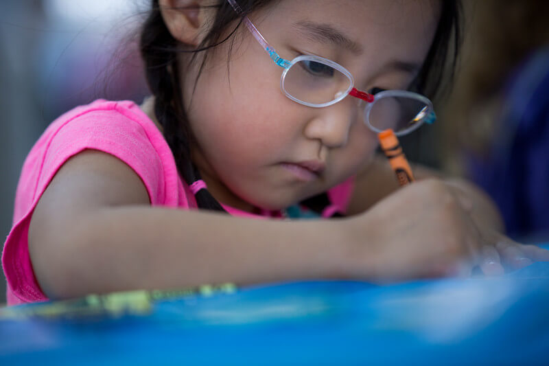 A child wearing glasses colors coloring with an orange crayon.