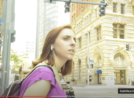 A still from the submitter's film that shows a teen with a purple shirt, a backpack and earbuds in her ears. She's in a city and looks like she's about to cross a street.