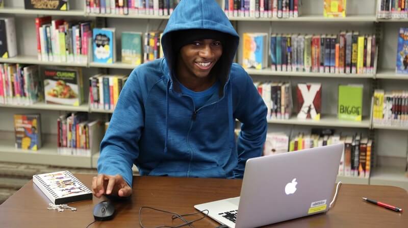 A smiling teen wearing a blue hoodie uses a laptop in front of a shelf of comic books