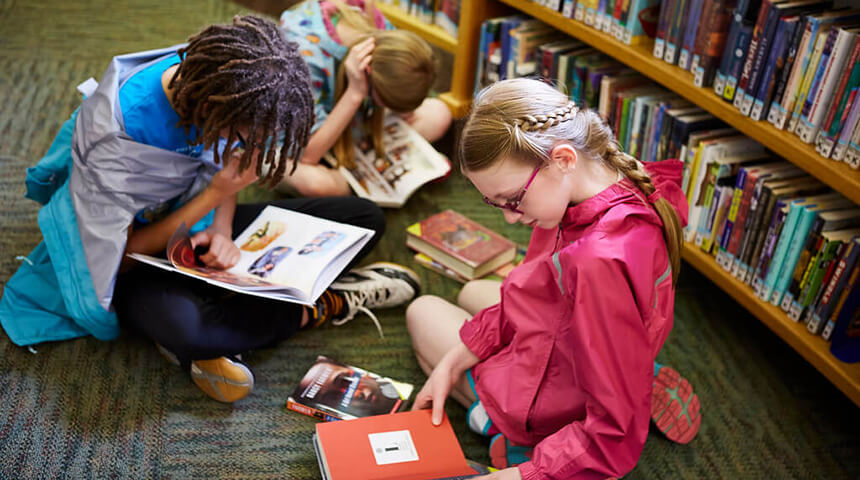 Three tweens sit on the floor of the library in front of a bookshelf, each reading a book