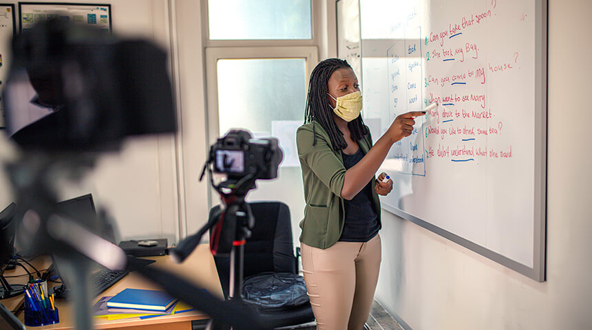 A teacher wearing a face mask writes on a white board in front of digital cameras on tripods