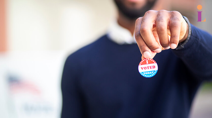 Adult wearing a blue sweater holds an "I voted" sticker in their hand