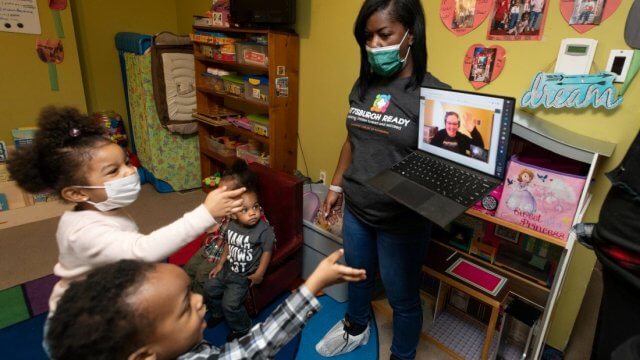 A teacher holds a laptop displaying a CLP virtual storytime to a group of young children