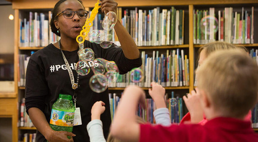 CLP children's librarian blowing bubbles through a yellow bubble wand for children at storytime.