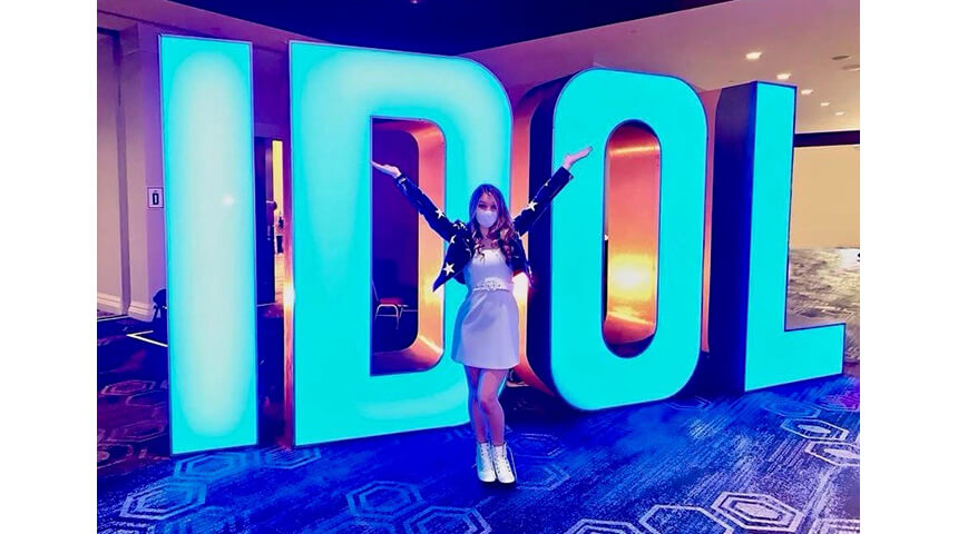 Teen girl in white dress and face mask stand in front of a glowing "American Idol" sign