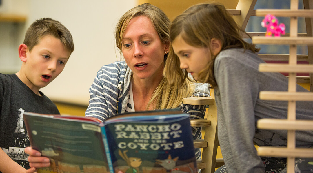 A caregiver and two children reading a book out loud in the children's room.