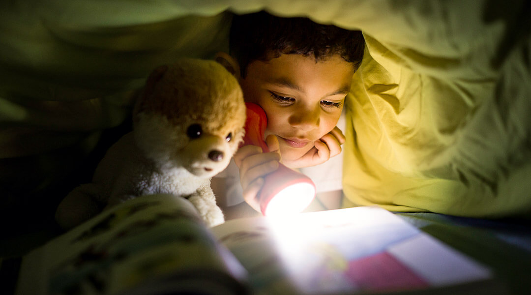 A child reading a book with a flashlight under the covers.