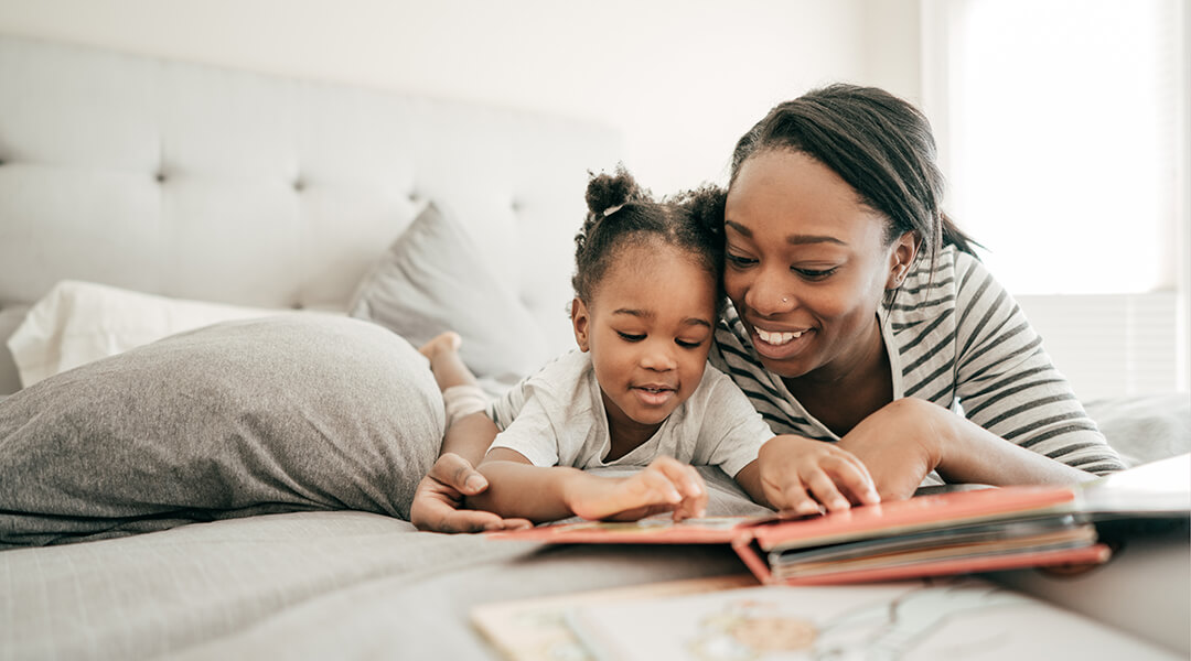 A smiling adult and toddler looking at a book together.