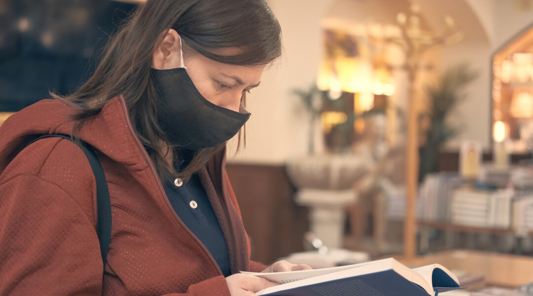 Person wearing a mask, coat and purse reading a book with tables of stacked books in the background.