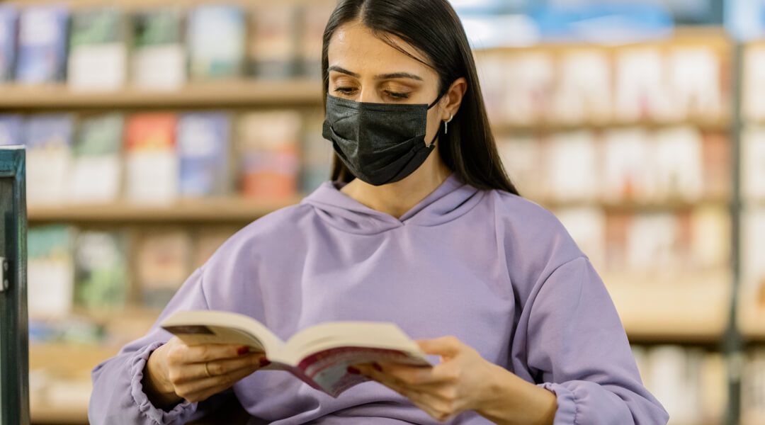Person with mask reading a book with book shelves in the background.