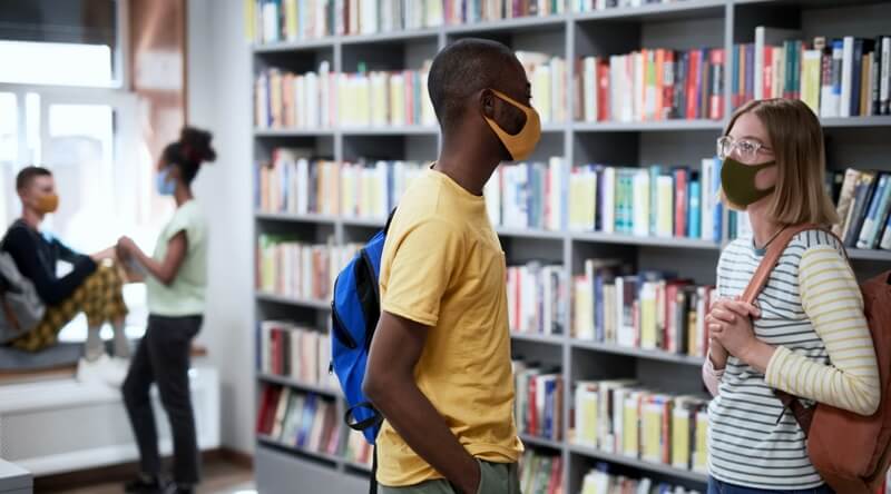 Teens with face masks conversing in a school library.