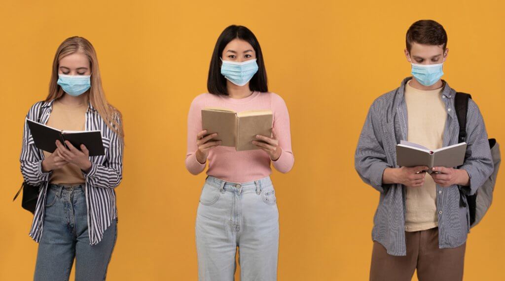 Three teens standing in a row wearing face masks and holding open books.