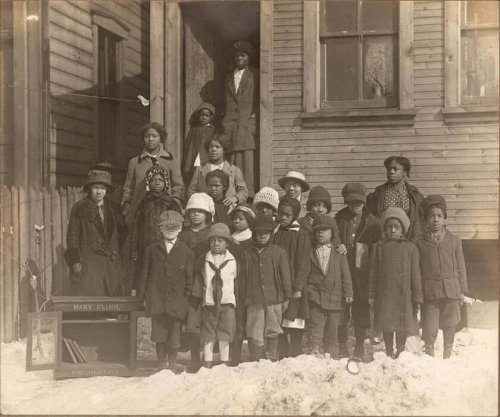 Old black and white photography of a group of children standing near a "home library" cabinet, c. 1928