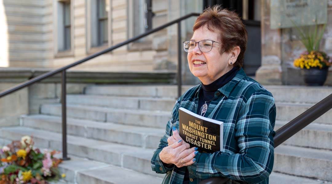 Mary Jane Kuffner Hirt on front steps of CLP-Main holding book titled "The Mount Washington Transit Tunnel Disaster."