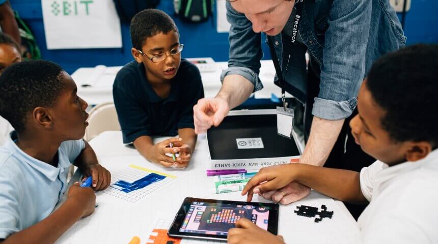 A group of teens work at a table with a library staff member on an iPad and other materials..