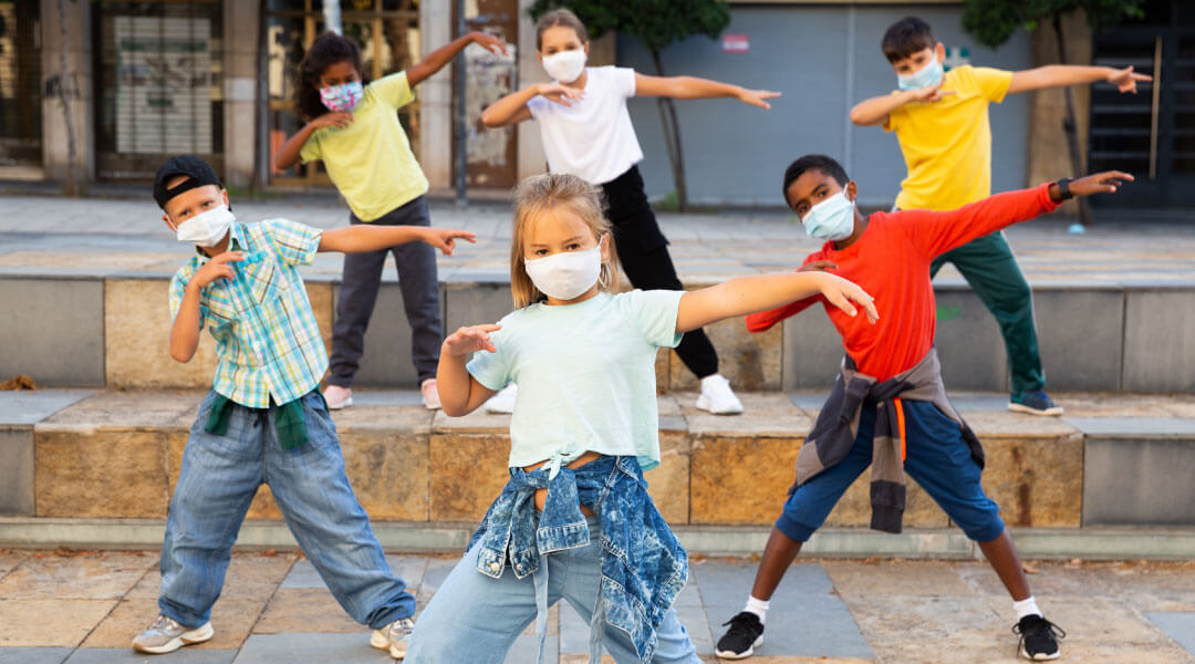 Young children posed in dance positions on building steps wearing face masks.