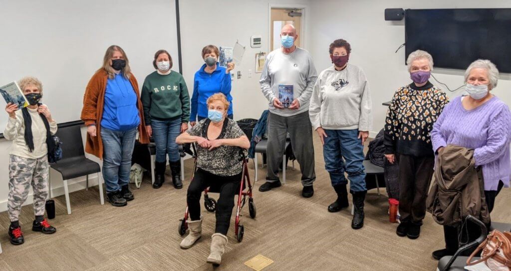 Masked people stand in a semi-circle. Two of them hold a book for a book club.