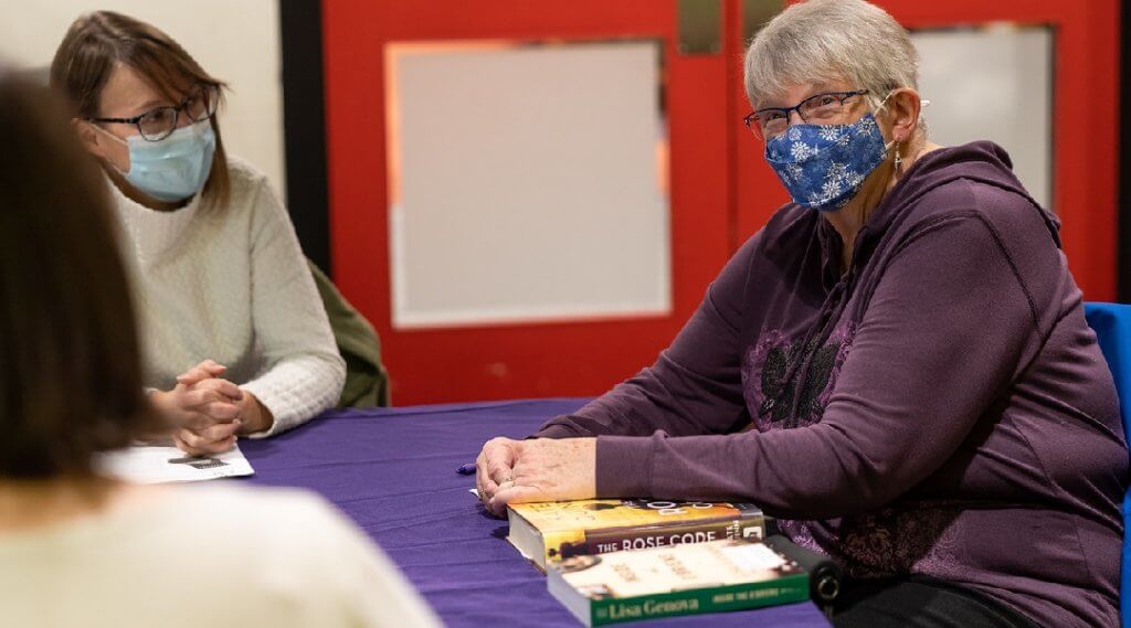 Two masked adults sit at a table with books by their folded hands