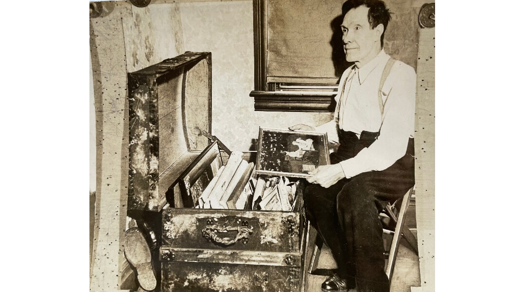 Black and white photo of John Kane, a middle aged white man, seated, looking at framed artwork in a trunk.