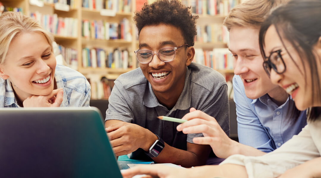 Four teens crowded around an open laptop in the library, smiling