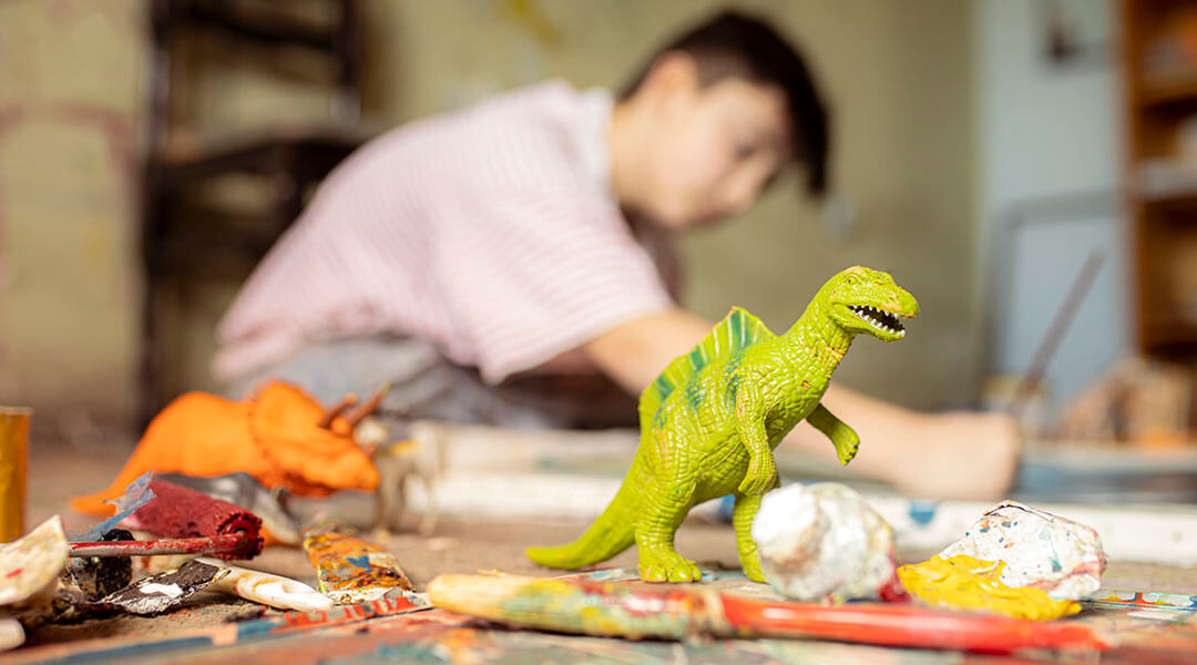 Dinosaur figures and art supplies on a table with a teen doing art in the background.