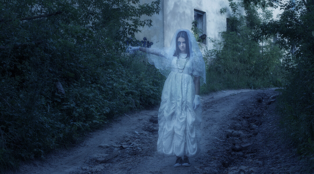 Young adult dressed in white dress and gloves on a dirt road, pointing toward an abandoned house.