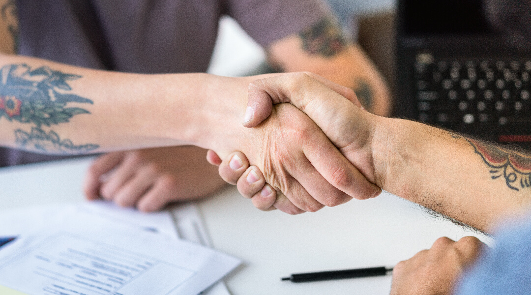 Close up of two adults with forearm tattoos shaking hands in an office.