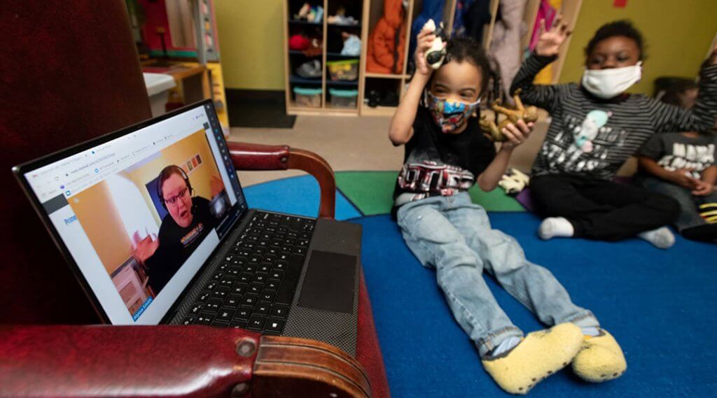 Several children sit on the floor of a classroom watching a librarian via video call.
