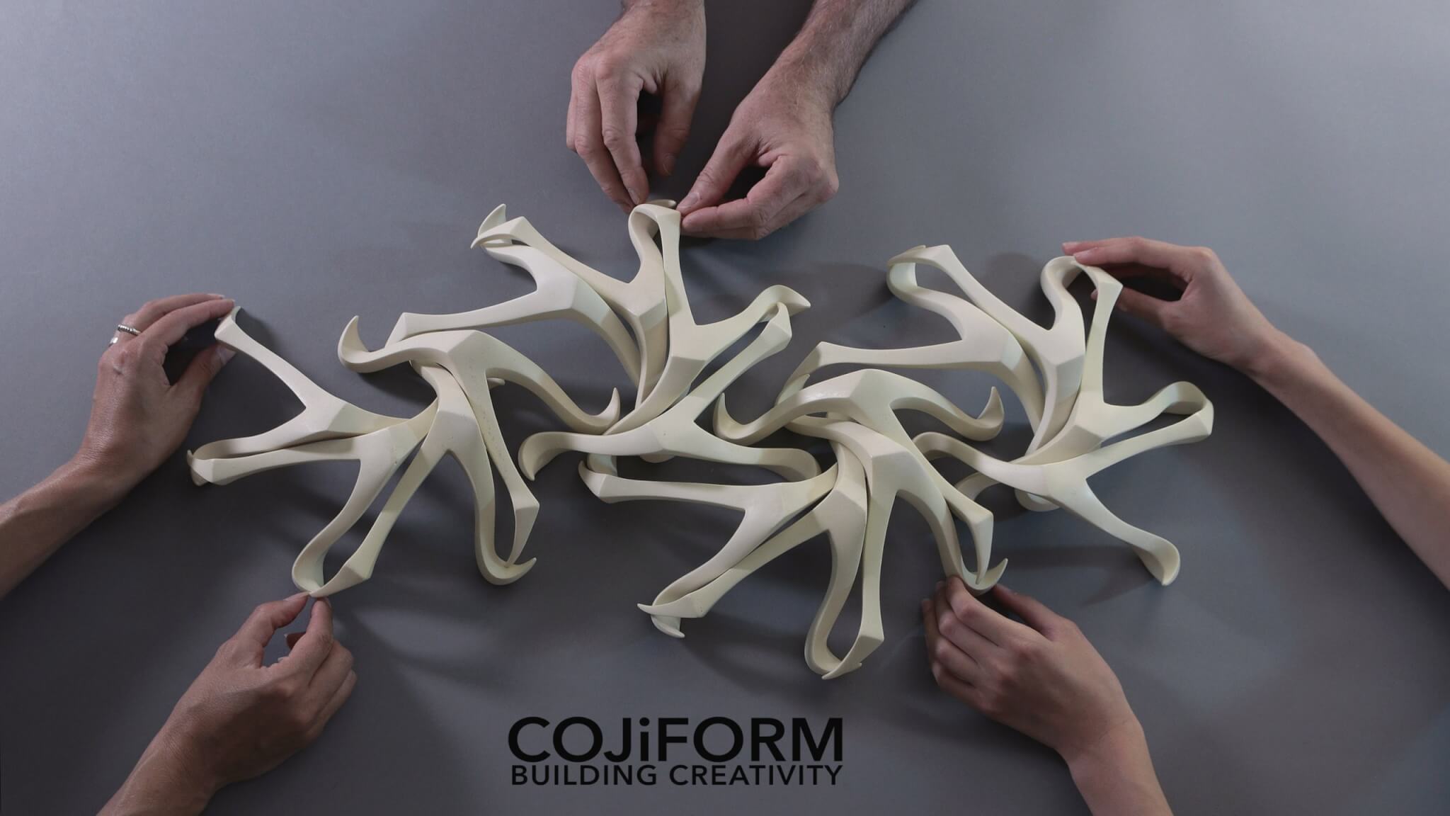 Three pairs of hands manipulating a cojiform sculpture on a table.