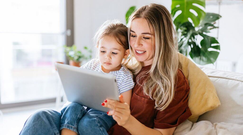 Mother and daughter using a digital tablet together. They are sitting on the sofa at home. The daughter is sitting on his mothers lap. Very hapy and smiling.