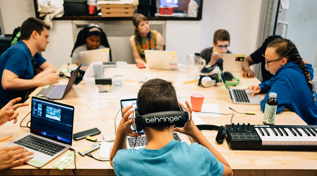 Teens sit around a table of laptops and sound mixing equipment in a music studio in the Teenspace.