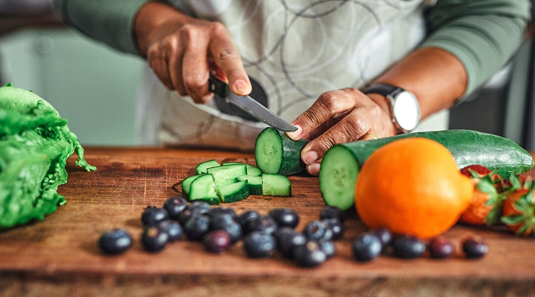 An adult slices zucchini on a cutting board with other vegetables.