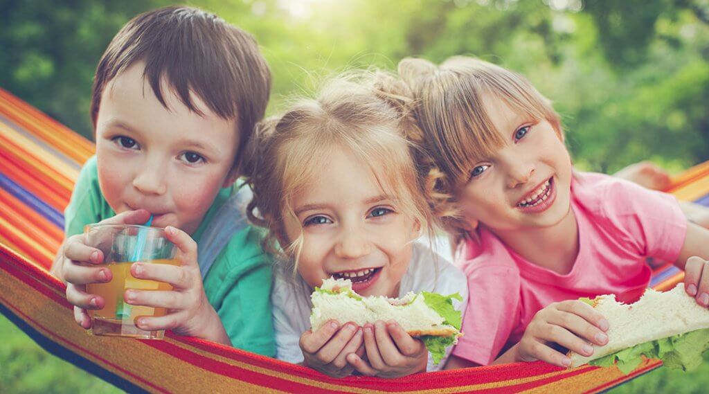 Three children eat snacks while lying on a hammock.