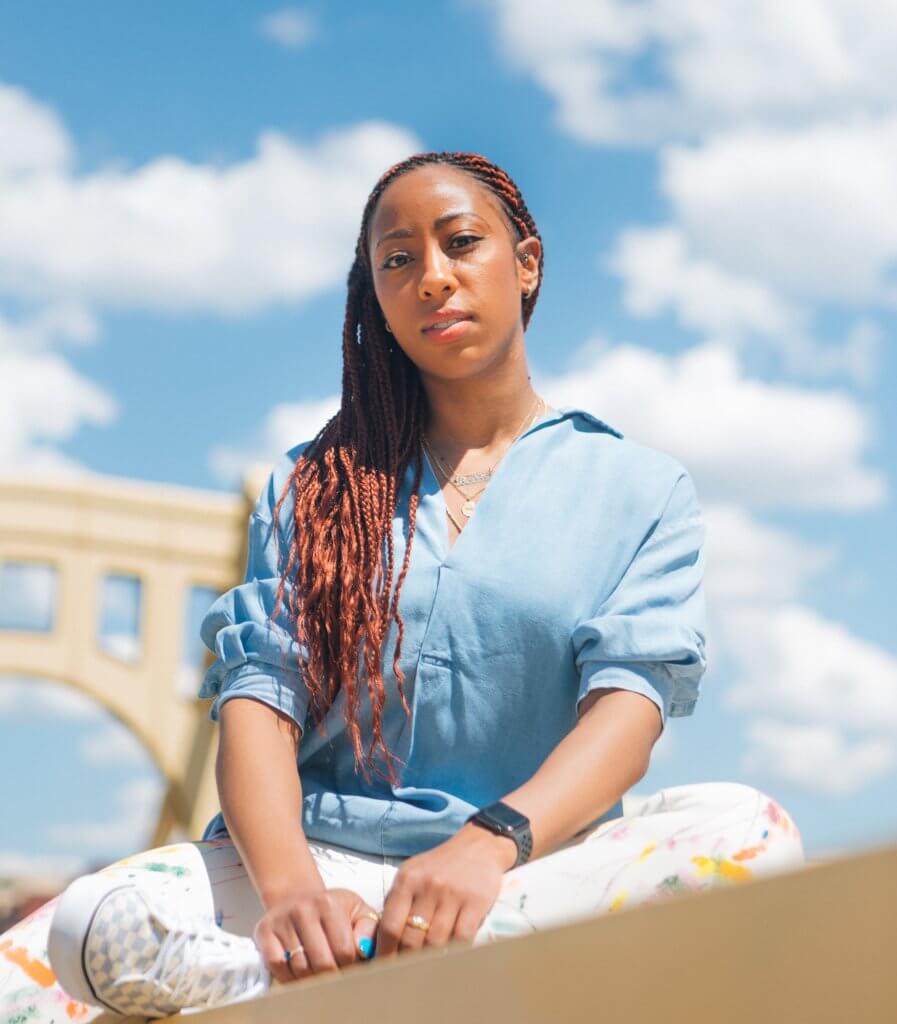 Artist Janel Young poses on one of Pittsburgh's yellow bridges in front of a cloudy blue sky. 