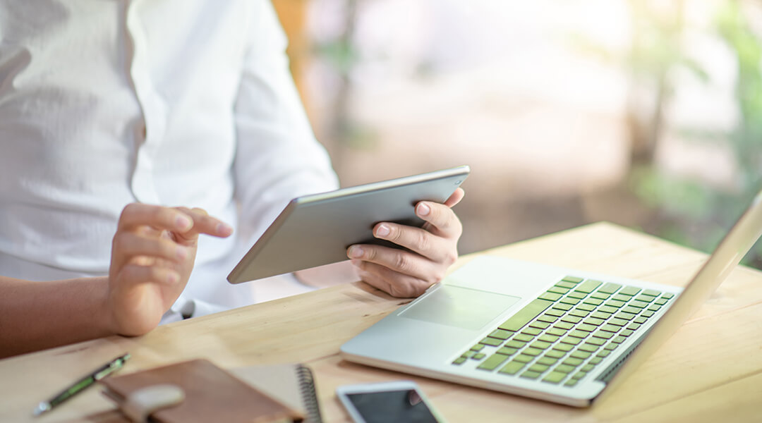 A person references a tablet while working at a laptop.