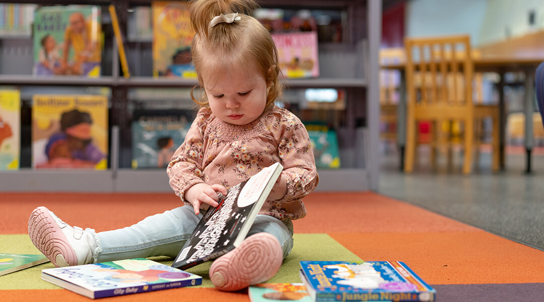 A baby sits on the floor in the library, playing with board books.