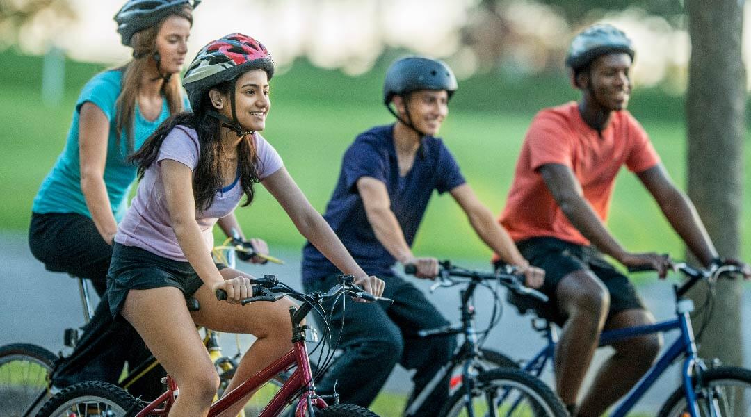 Four teens ride bikes together, wearing helmets.