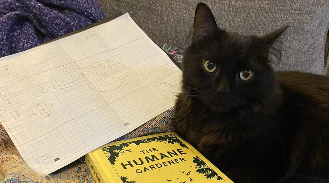 Black cat sitting with a book titled The Humane Gardener and a piece of graph paper