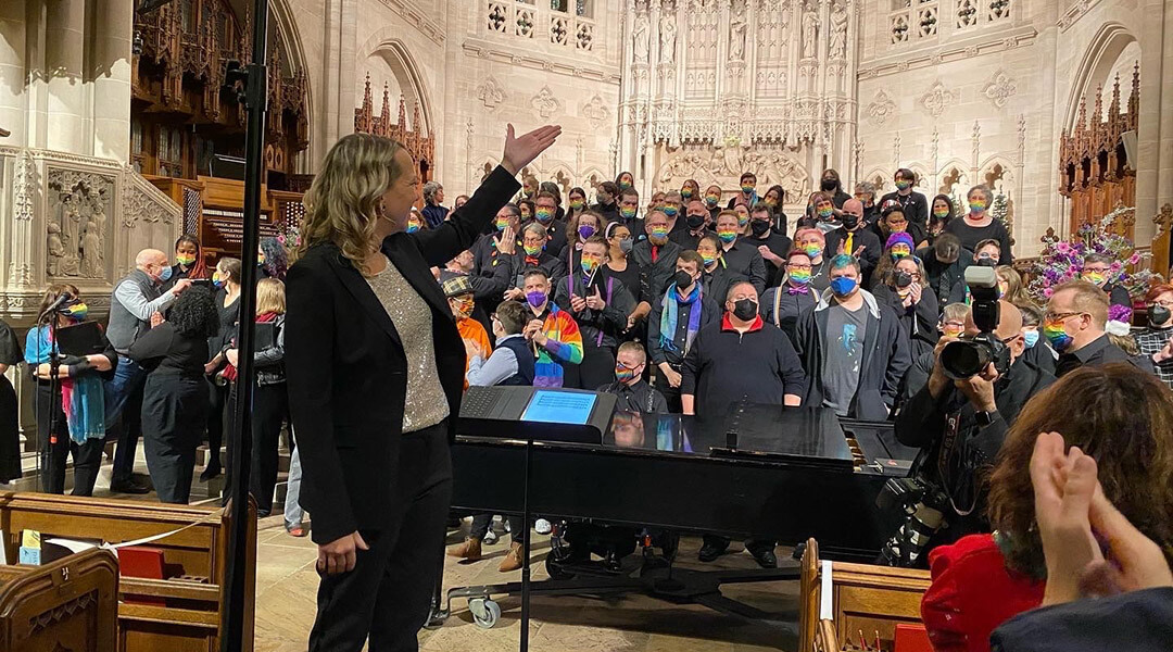 A choir director stands in front of a group of singers wearing rainbow masks.