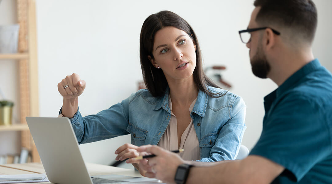 An adult gestures as they explain a concept to another person sitting at a laptop.