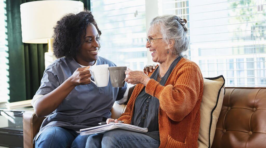 A senior in their 80s sitting on a couch, having coffee and conversing with a younger adult.